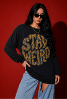 A woman modeling a black long-sleeve graphic T-shirt with a large leopard-print 'STAY WEIRD' design, paired with light blue jeans, against a solid red studio background.