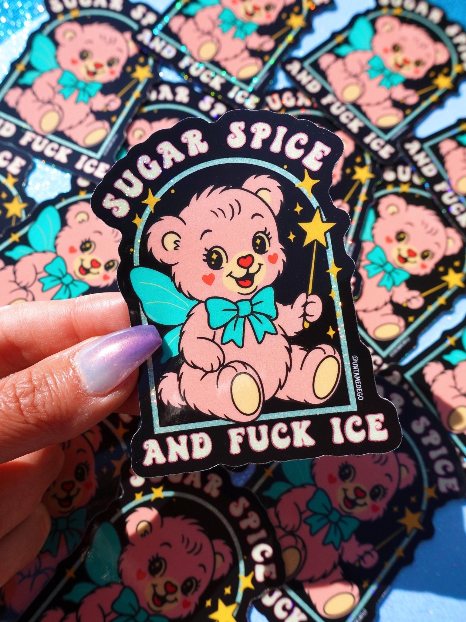 Holographic glitter protest sticker reading "Sugar Spice and Fuck ICE" held by a hand with painted nails, surrounded by multiple copies of the same sticker scattered on a dark surface.