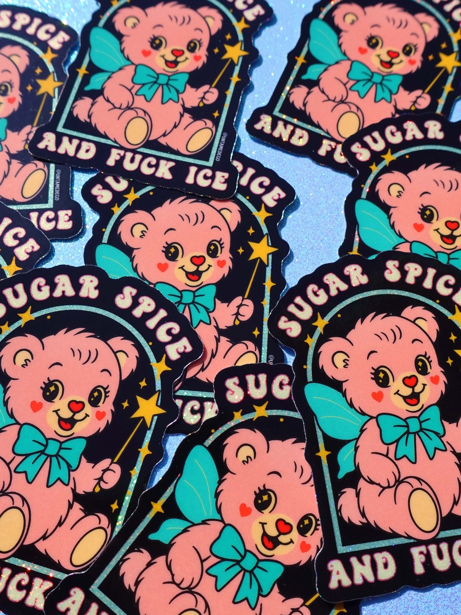Holographic glitter protest sticker reading "Sugar Spice and Fuck ICE" held by a hand with painted nails, surrounded by multiple copies of the same sticker scattered on a dark surface.
