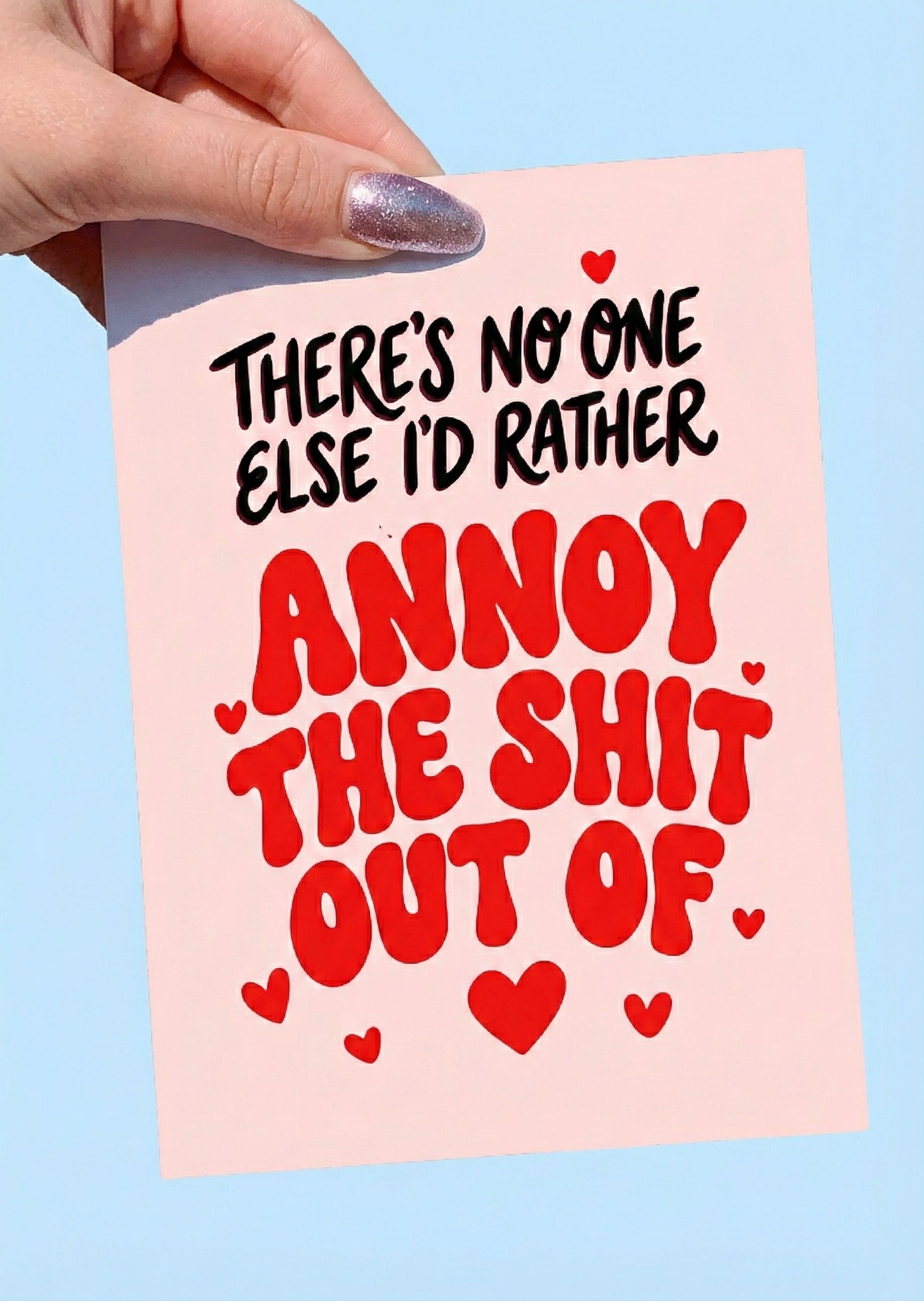 Greeting card with bold red and black text and heart graphics on a pink background, photographed being held by a hand
