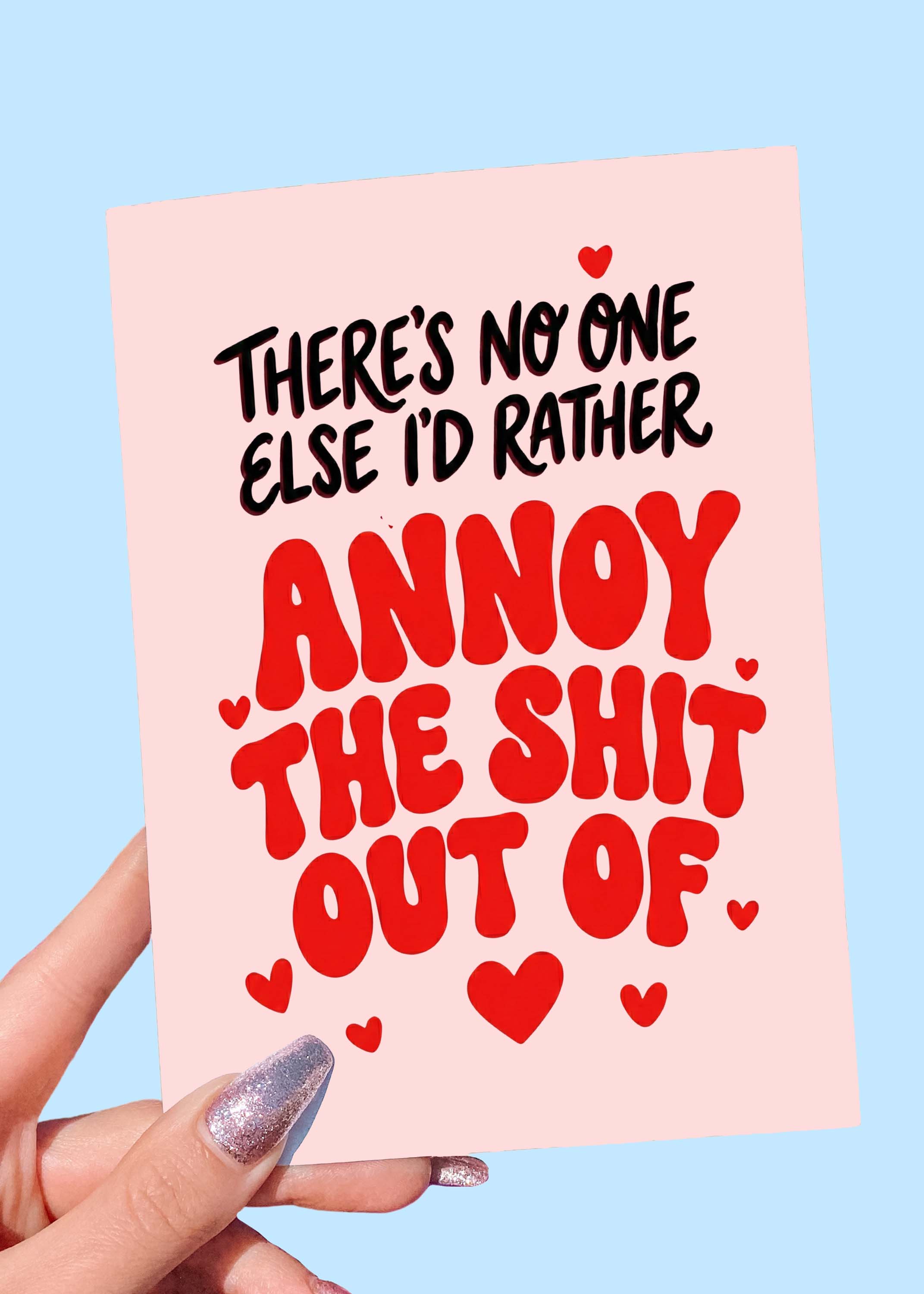 Greeting card with bold red and black text and heart graphics on a pink background, photographed being held by a hand
