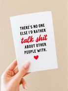 Greeting card held in a hand against a neutral background. The card has a white background with bold black text that reads, “There’s no one else I’d rather,” followed by the phrase “talk shit” in red script-style lettering, and then black text reading, “about other people with.” A small red heart icon appears centered near the bottom of the card. The card is vertically oriented and blank inside