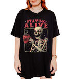 Black oversized graphic tee with red text reading "STAYING ALIVE" above a skeleton holding a steaming red coffee mug labeled "Coffee," with sparkle accents and a rectangular border.