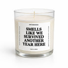 A single-wick candle in a clear glass jar with cream-colored wax and a white label featuring bold black text that reads 'SMELLS LIKE WE SURVIVED ANOTHER YEAR HERE' by Untamedgo, shown on a clean white background.