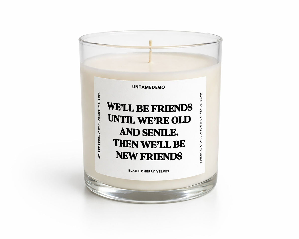 A white scented candle in a clear glass jar with a white label featuring black text that reads, 'UNTAMEDEGO WE'LL BE FRIENDS UNTIL WE'RE OLD AND SENILE. THEN WE'LL BE NEW FRIENDS.